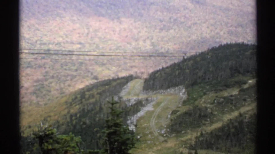 1971 Vermont 8mm frame of a mountain road and evergreen foreground against a broad autumn hillside crossed by utility lines.