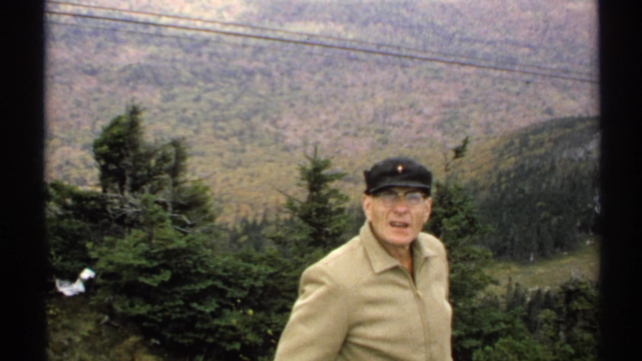 Elderly man in a beige jacket and black cap stands before evergreen trees and a wooded mountain valley.