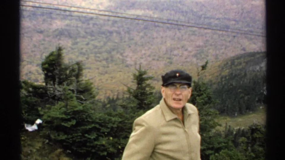 In 1971 Vermont home-movie footage, an elderly man faces the camera near a mountain overlook with evergreen trees, power lines, and a forested valley behind him.