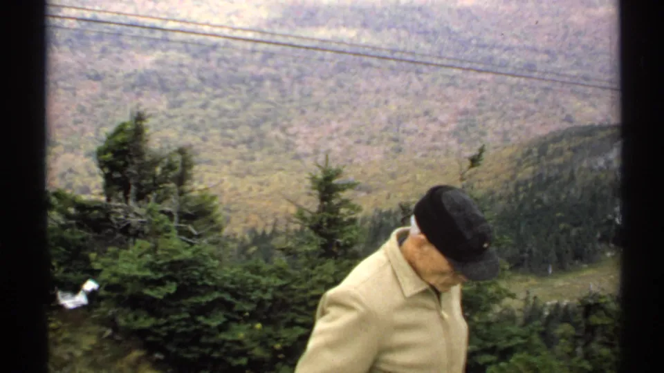 The elderly man lowers his gaze beside evergreen trees, with overhead power lines and a wooded Vermont mountain valley visible beyond him.