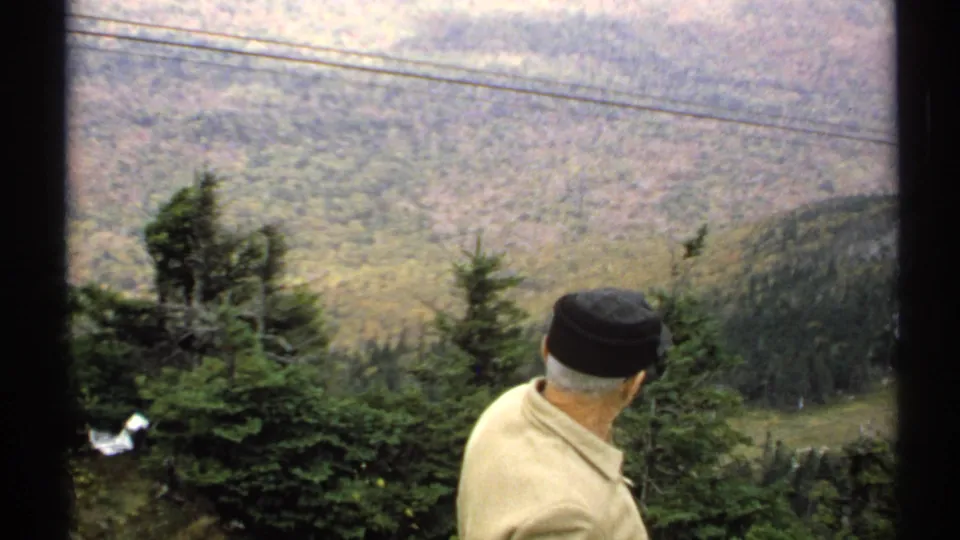 Seen from behind in 1971 Vermont footage, the man turns away from the camera to look across the muted autumn valley and surrounding evergreens.
