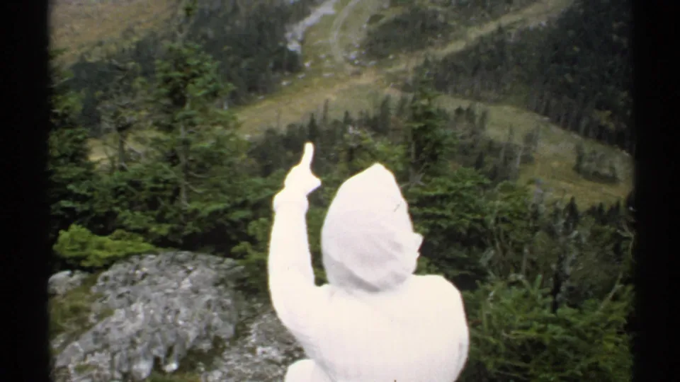 In the Vermont overlook scene, the white hooded visitor raises one hand while facing the valley and distant winding path.