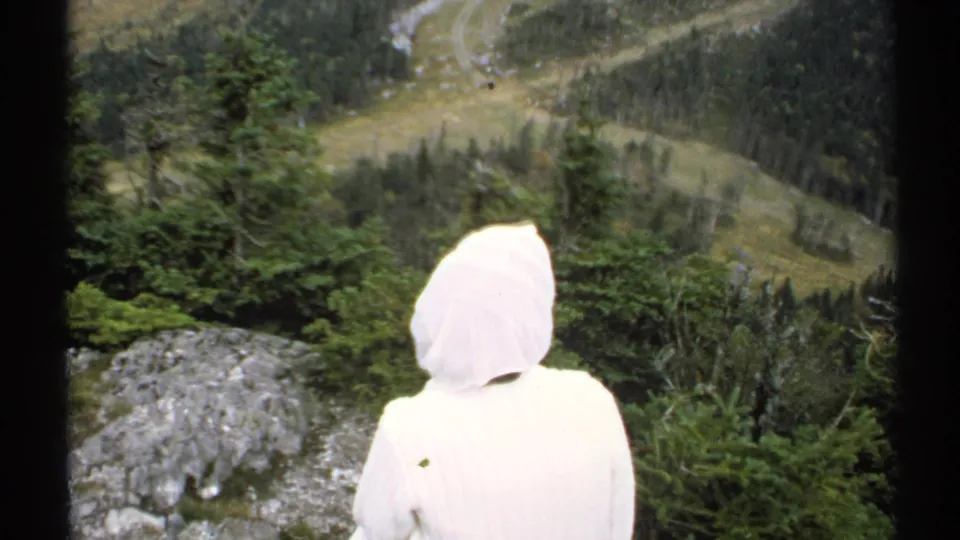 A white hooded figure, seen from behind in 1971 Vermont footage, looks over evergreens, rock, and a broad wooded valley.