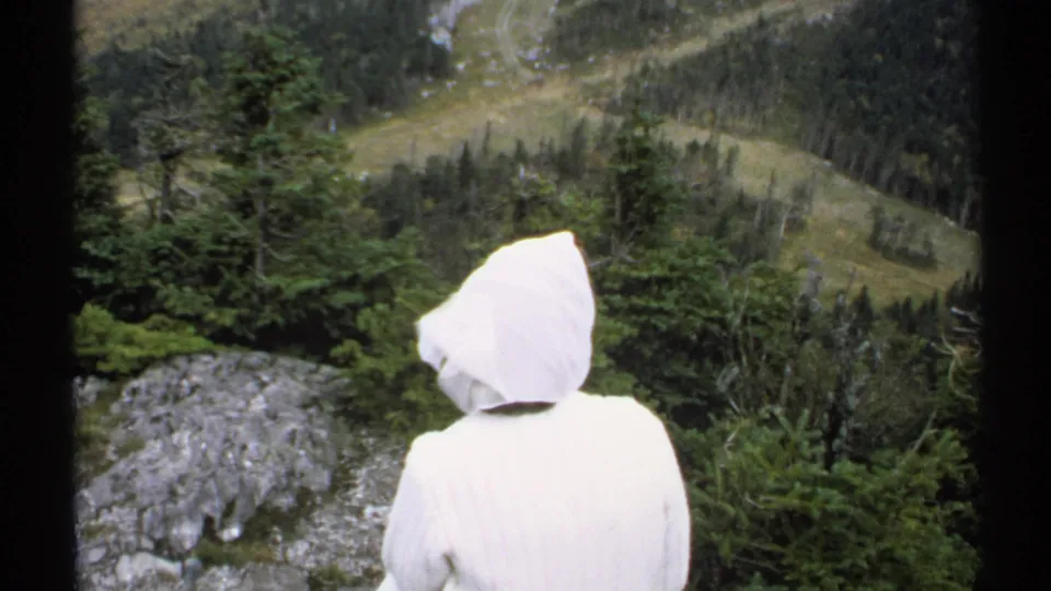 A hooded figure stands with their back to the camera at a Vermont mountain overlook, with trees, rock, and distant hillside beyond.