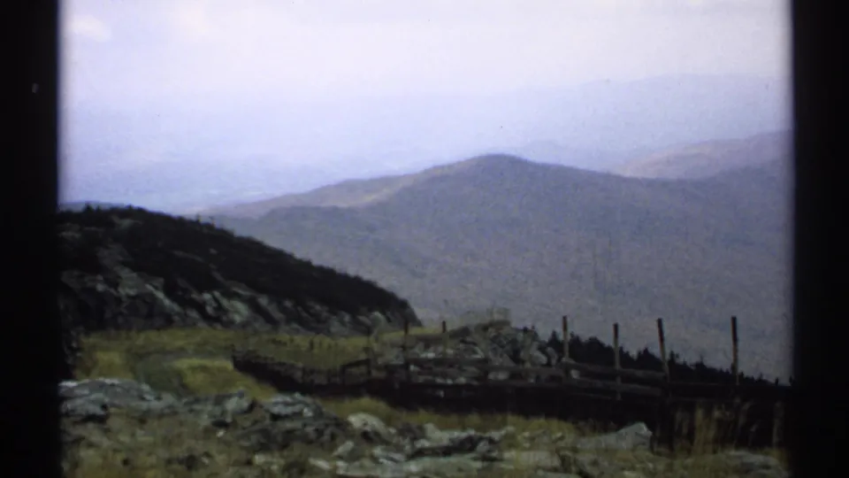A Vermont overlook view shows a wooden fence, rocky ground, dark treeline, and hazy mountain ridges in the distance.