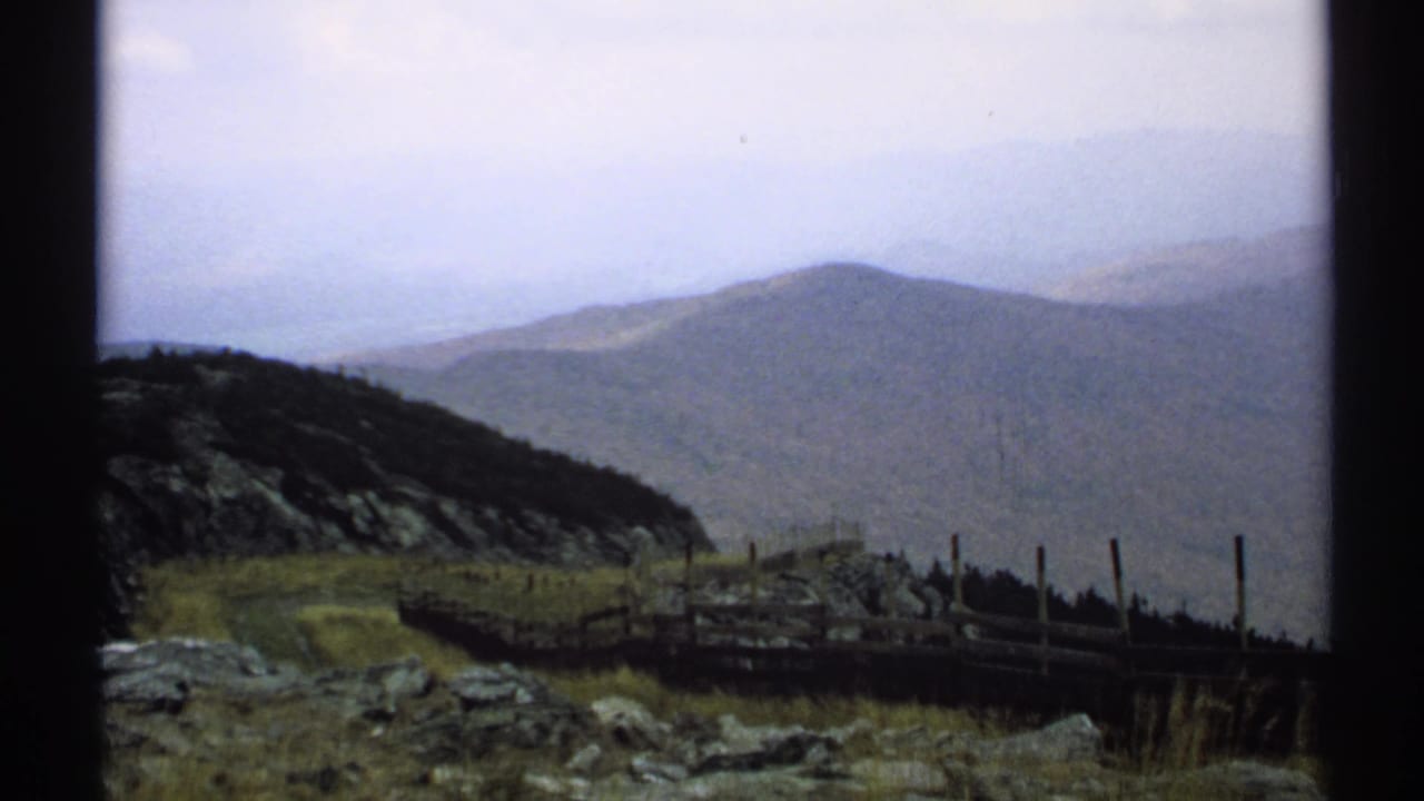 Hazy mountain ridge beyond rocks, grass, and a wooden fence at a high overlook.