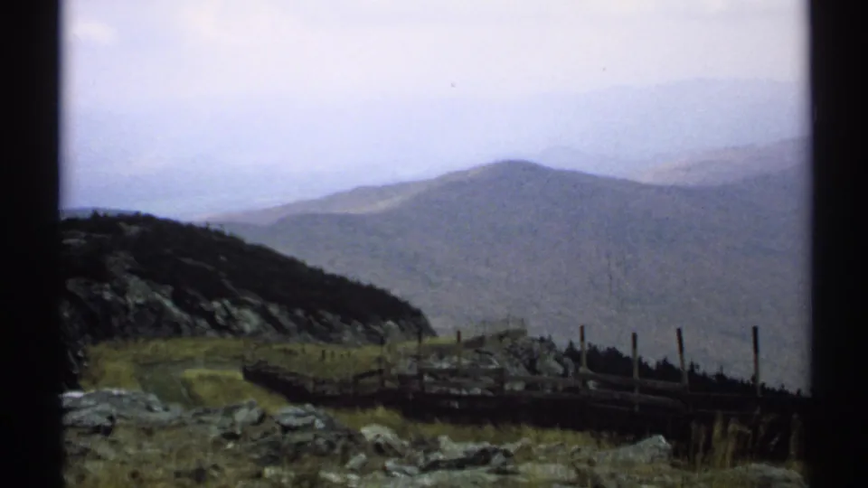 1971 Vermont 8mm frame showing a hazy mountain vista with rocky foreground, grasses, a wooden fence, and layered ridges.
