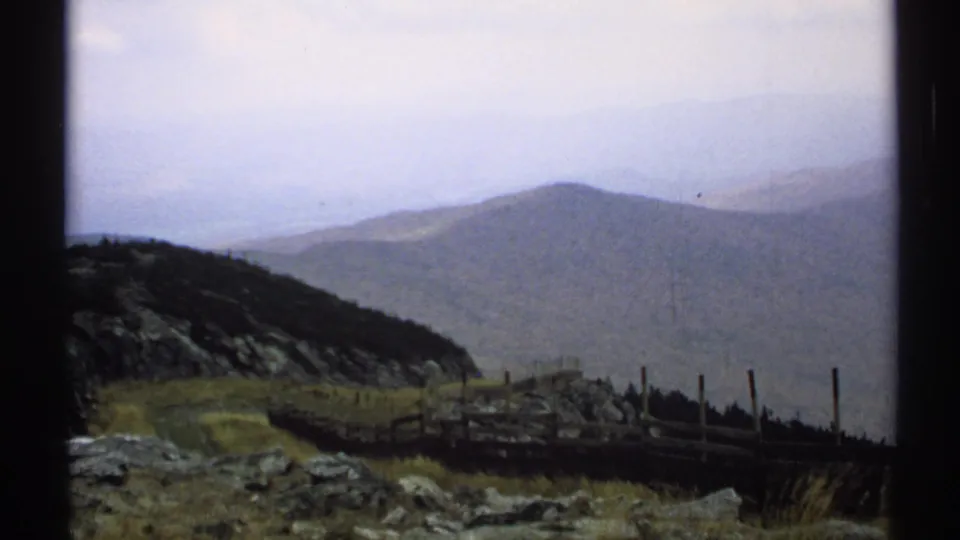 1971 Vermont home-movie view of layered ridges under haze, with fence posts, rocks, and a dark slope in the foreground.