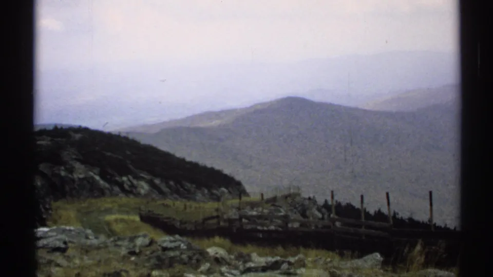 1971 Vermont 8mm landscape frame with rocks, grasses, a rough fence, and distant mountain ridges softened by haze.