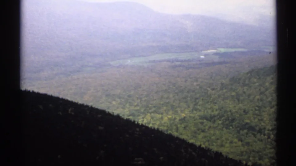 1971 Vermont 8mm frame looking across a forested valley toward hazy ridges, with a small pale water or clearing visible below.