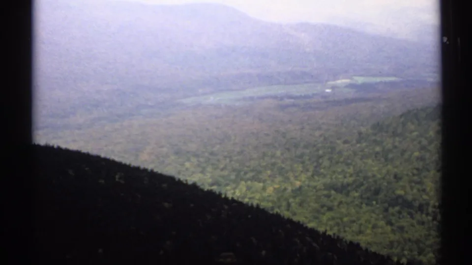 1971 Vermont 8mm frame showing an elevated view across dense green forest toward hazy mountain ridges and a distant lake or valley.