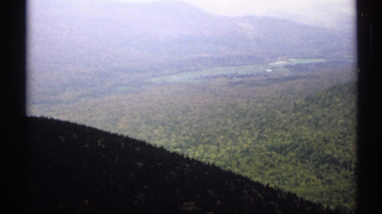 1971 Vermont 8mm frame with dark vertical edges around a hazy elevated view of forested hills, distant ridges, and a pale lake or valley.