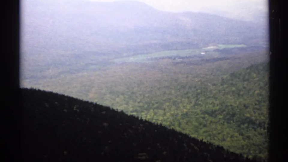 1971 Vermont 8mm frame with dark vertical edges around a hazy elevated view of forested hills, distant ridges, and a pale lake or valley.