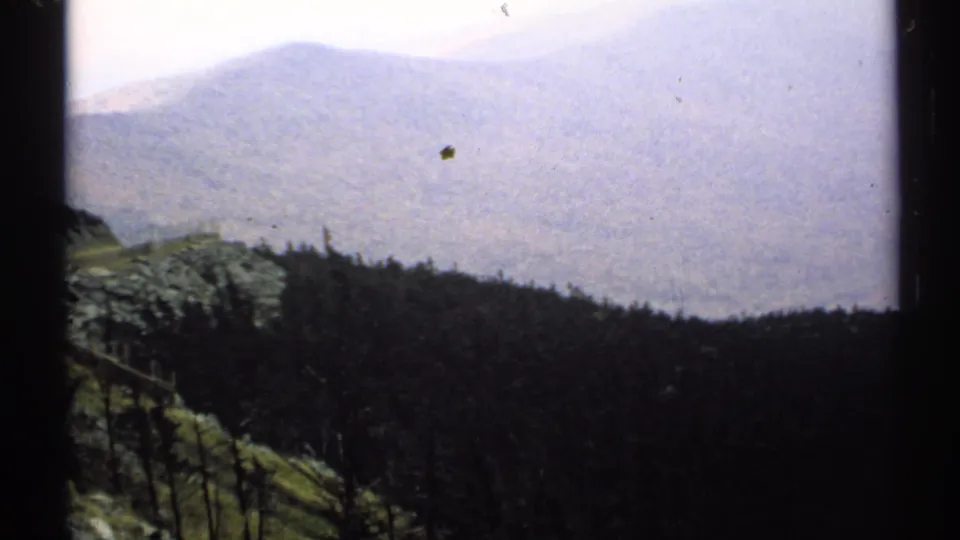 1971 Vermont 8mm frame looking from a high rocky foreground past a dark treeline toward a soft, hazy mountain ridge.