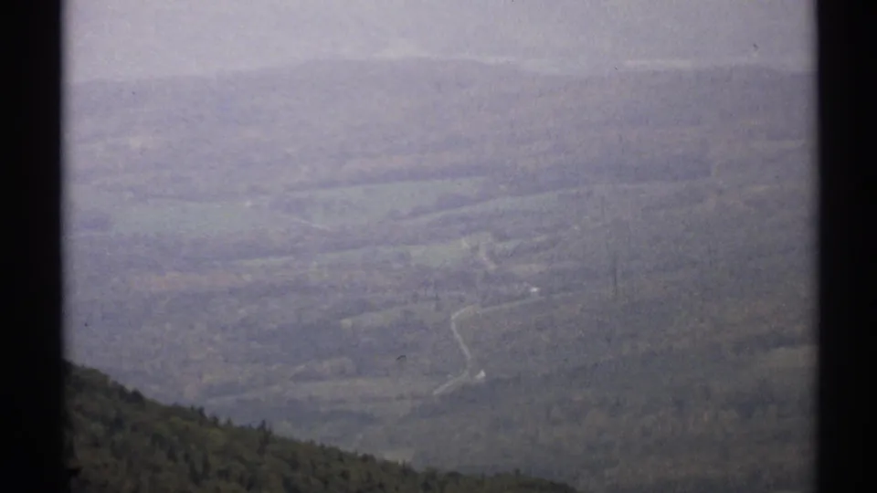 1971 Vermont home-movie frame with a soft, hazy view of forested hills and a road tracing through the valley.