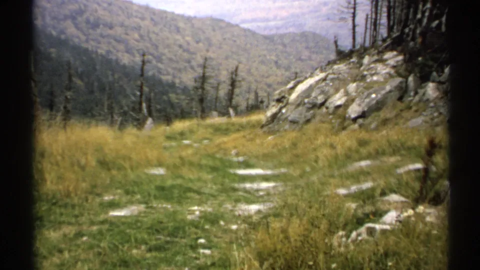 1971 Vermont home-movie frame of a rocky grassy trail passing along a mountainside, with sparse trees and layered forested hills beyond.