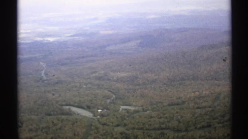 1971 Vermont 8mm view from a high vantage point over forest, winding waterways, small structures, and hazy hills.