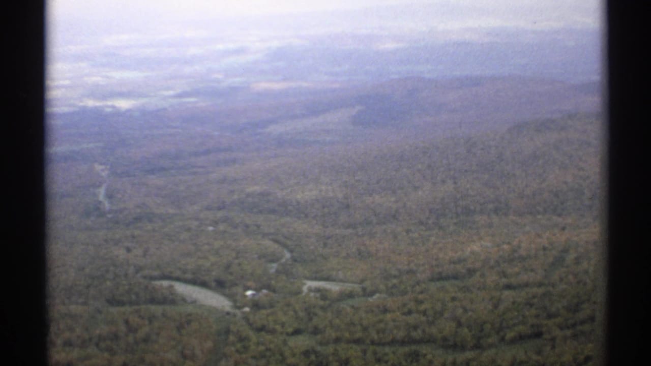 Wide aerial view across wooded hills, curving roads, and distant hazy ridges.