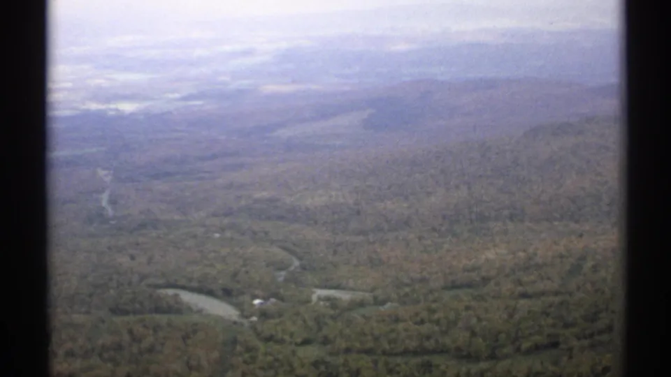 1971 Vermont aerial home-movie frame showing forested hills, a pale winding road, scattered buildings, and distant ridgelines through haze.