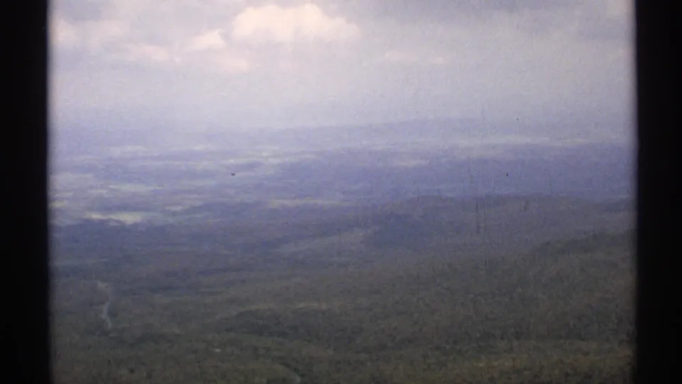 1971 Vermont 8mm frame looking across a hazy valley, with forested slopes, distant hills, and a winding road or stream near the lower edge.
