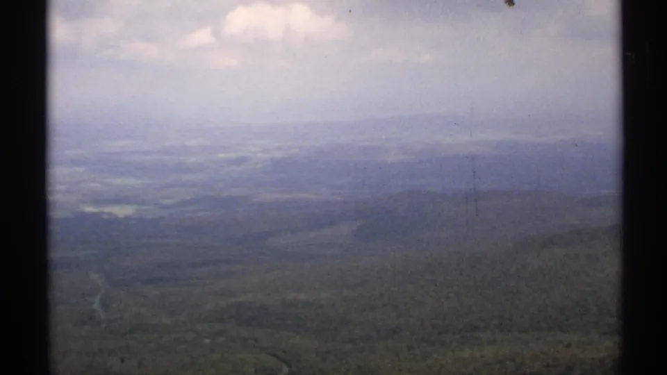1971 Vermont aerial view over a hazy forested valley, with rolling hills, a pale road or stream, and soft clouded light in the distance.