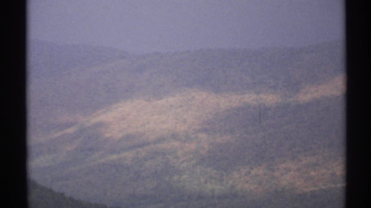 Soft aerial view of rolling wooded hills with pale patches and haze.