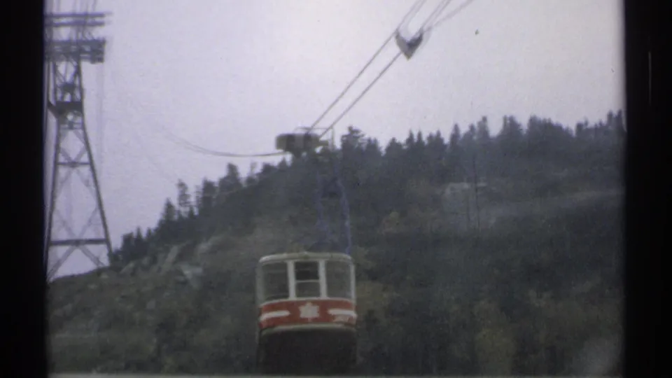Wider 1971 Vermont view of a red and white tram car ascending near a metal support tower and wooded ridgeline.
