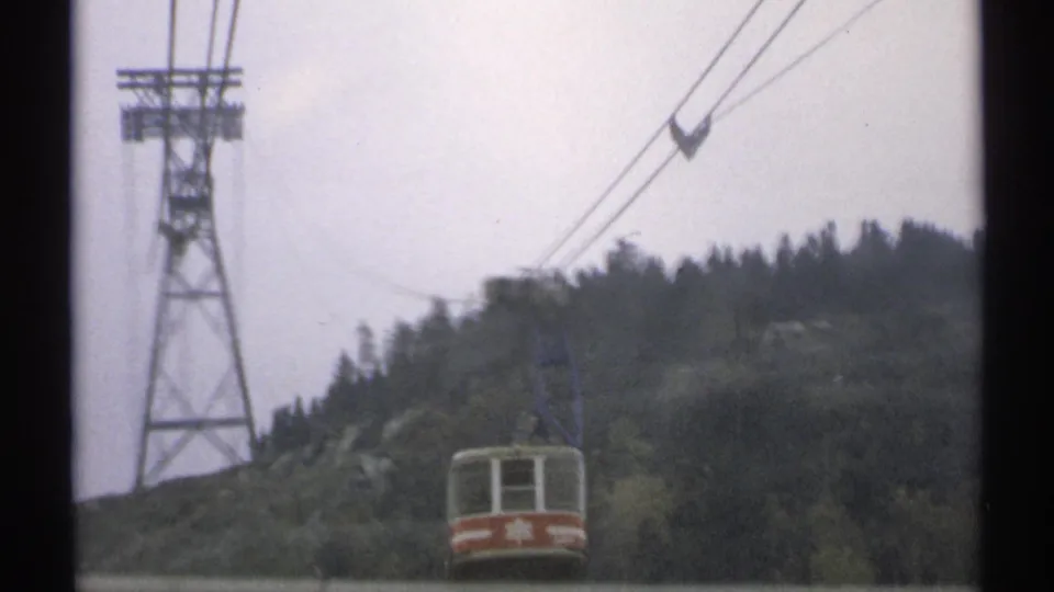 Distant red and white tram car in Vermont, framed with overhead cables, a support tower, and dark evergreen hillside.