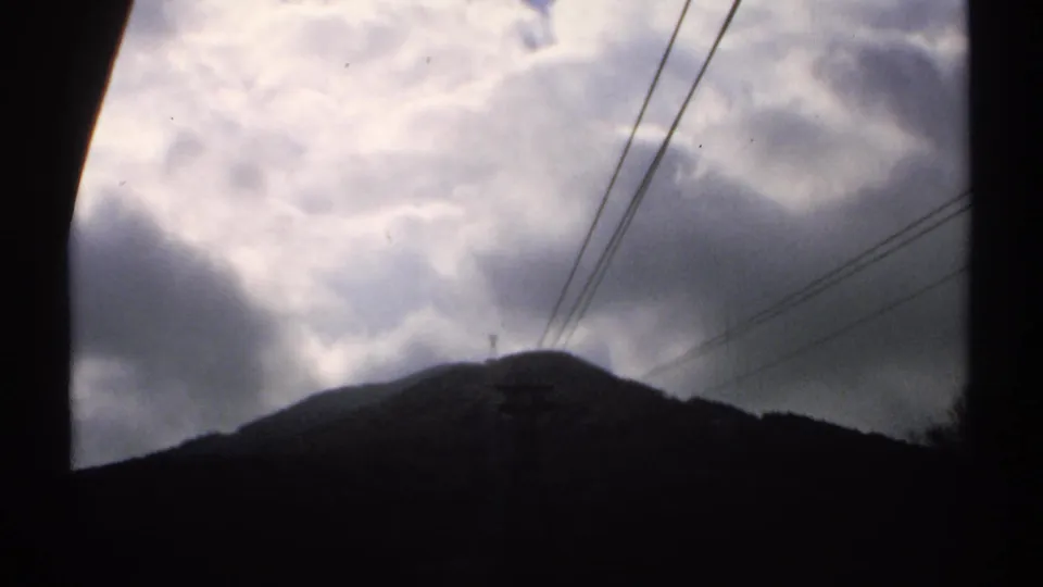 Vintage Vermont home-movie frame with brooding clouds above a dark ridge and visible utility infrastructure in the foreground.