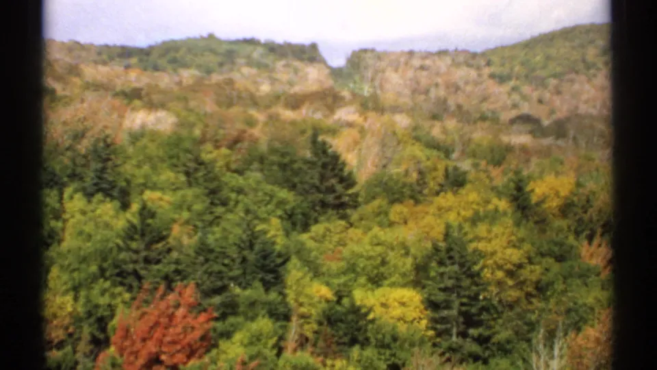 1971 Vermont 8mm frame of a brighter autumn forest with green, yellow, and red foliage below rocky wooded hillsides.