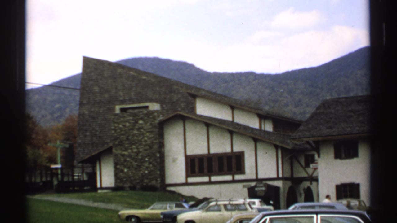 Chalet-style building with parked cars and forested mountains behind it.