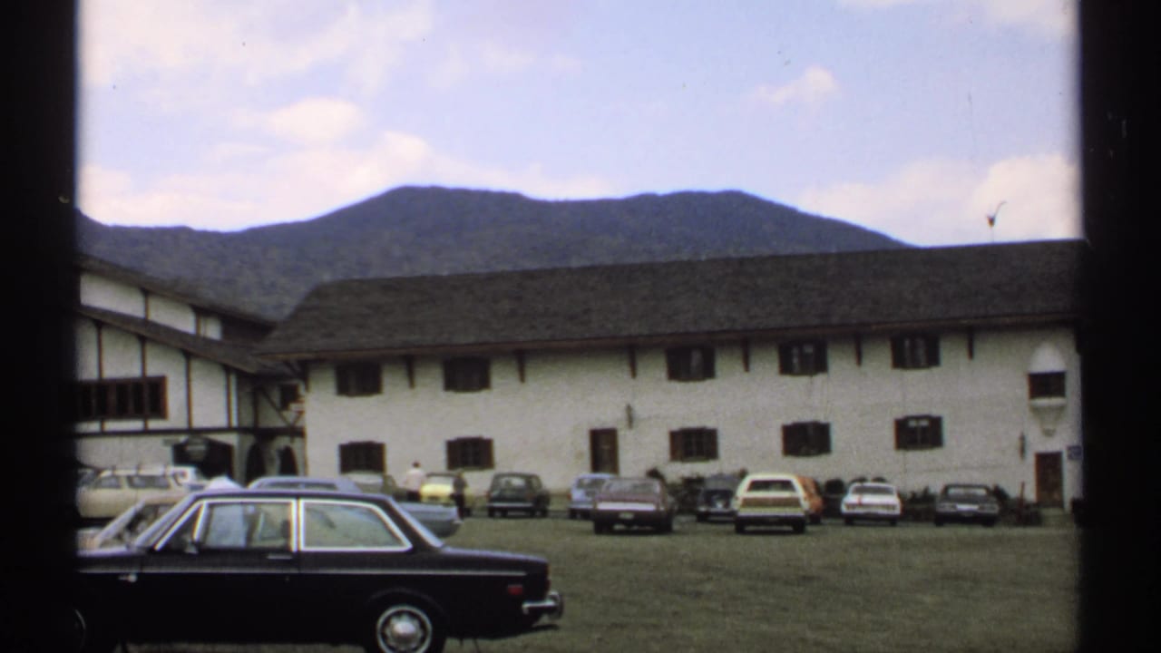 White and brown building with parked cars in front and mountains behind.