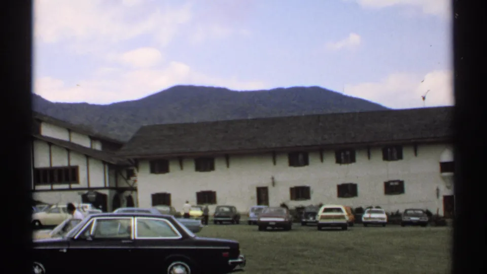 1971 Vermont frame with a black sedan in the foreground, a long white-and-brown building, other parked cars, and a wooded mountain backdrop.