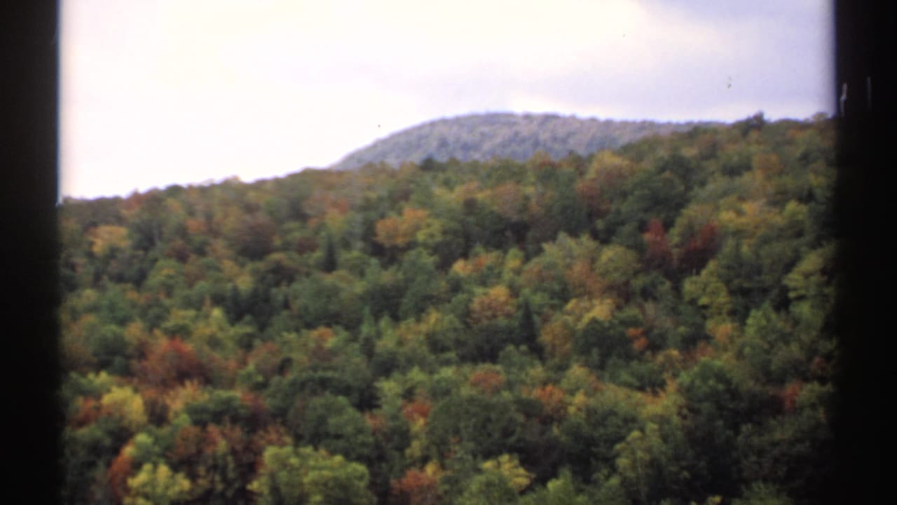 1971 Vermont home-movie frame with a rounded wooded hill and mixed fall foliage visible through dark 8mm frame edges.