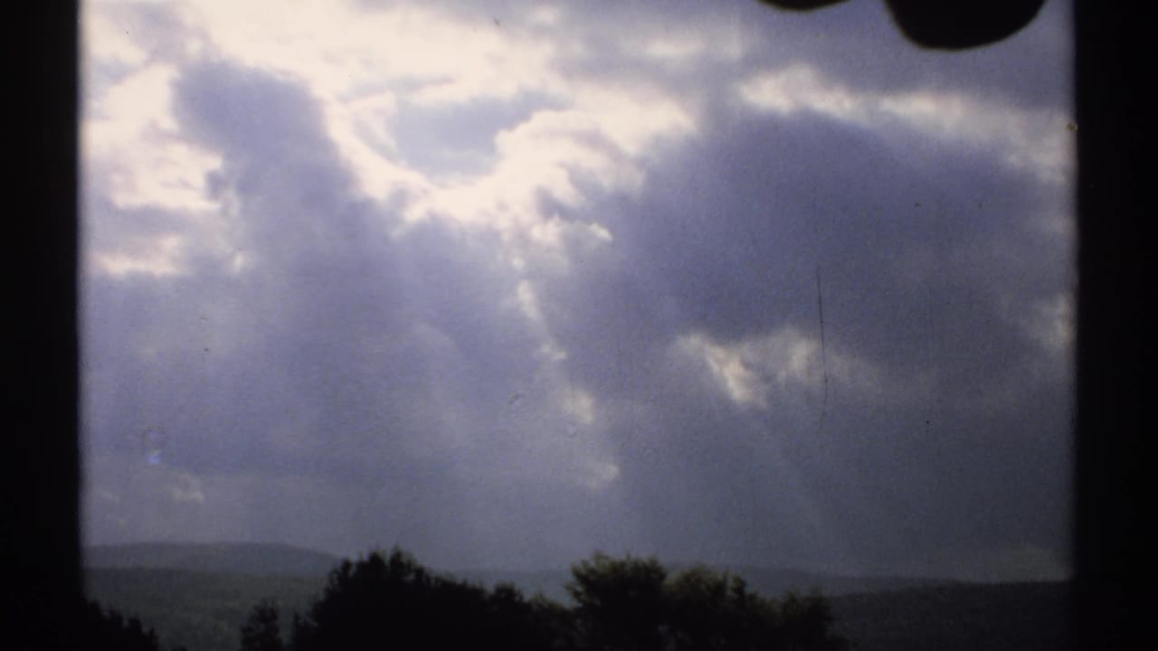 Dramatic cloudy sky over dark trees and distant hills seen through a window frame.