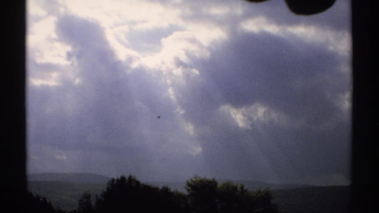 Cloud-filled sky with bright breaks above a dark rural treeline and low hills.