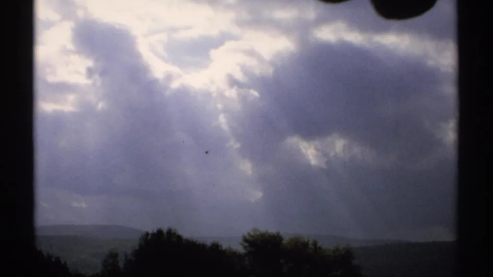 1971 Vermont home-movie view of layered clouds, filtered sunlight, distant hills, and a dark treeline through a window-like frame.