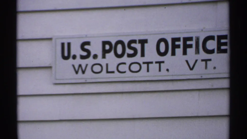 1971 Vermont 8mm frame showing a U.S. Post Office sign for Wolcott, VT mounted on white clapboard siding.