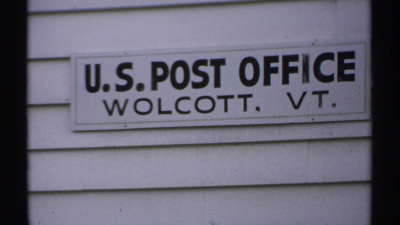 Close view of a U.S. Post Office sign reading Wolcott, VT on white clapboard siding.