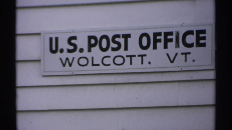 1971 Vermont 8mm frame showing a U.S. Post Office sign for Wolcott, VT mounted on a white clapboard building.