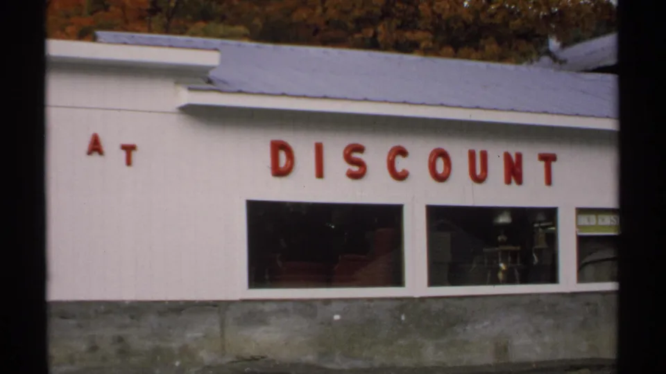 1971 Vermont home-movie frame of a white storefront section reading At Discount above dark display windows.
