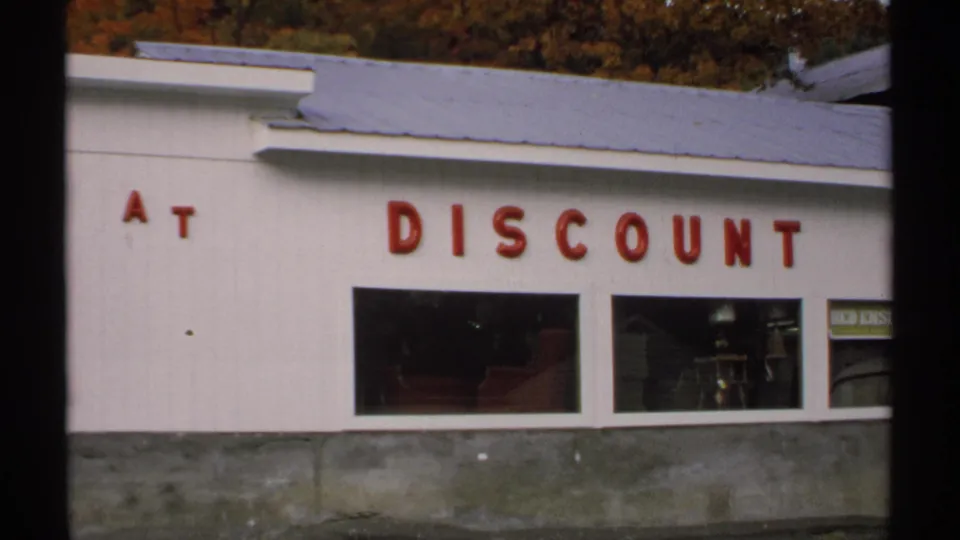 1971 Vermont home-movie frame showing part of a white retail storefront with red discount lettering and two display windows.