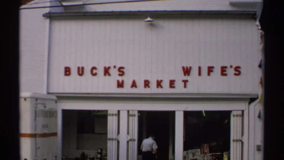 1971 Vermont frame of Buck's Wife's Market, with red lettering on white siding, open doors, and an adult entering the shop.