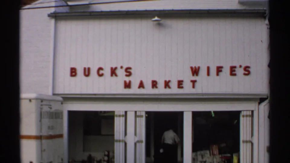 1971 Vermont storefront view of Buck's Wife's Market, showing red wall letters, open entry doors, and a man moving inside.