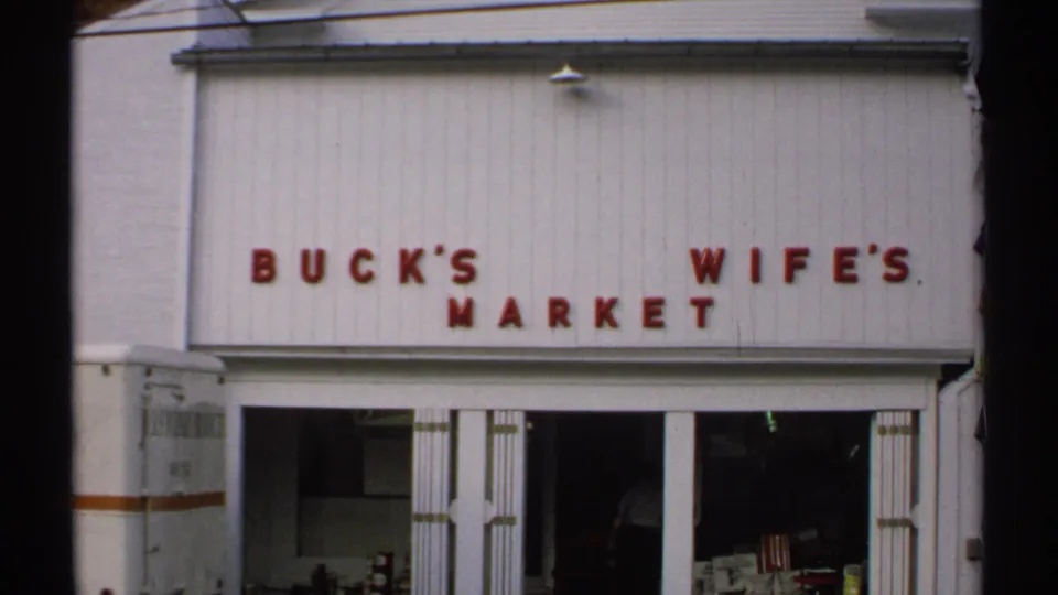 1971 Vermont home-movie frame holding on Buck's Wife's Market, with red signage, white facade, open doors, and a dark interior figure.