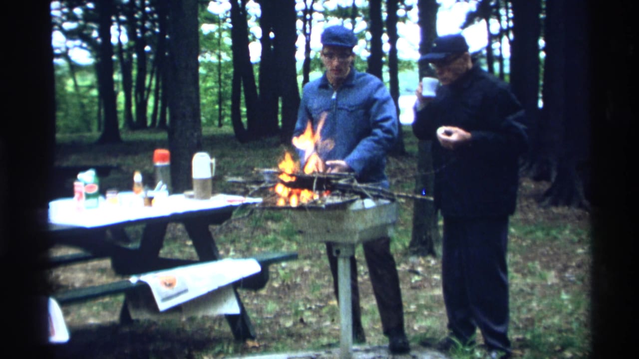 Two men stand beside a small campfire and picnic table in a wooded area.