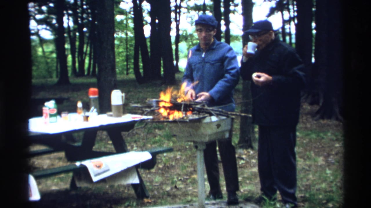 A man tends a campfire while another man drinks from a cup near a picnic table.