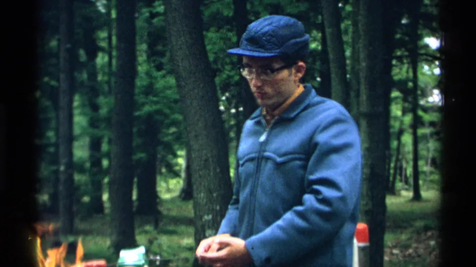 Closer 1971 Wisconsin 8mm view of an adult man in a blue jacket and cap near flames, with trees and picnic supplies behind him.