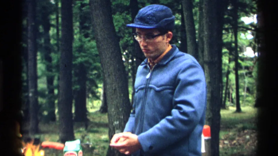 1971 Wisconsin home-movie frame of a man in a blue jacket and cap beside a campfire, surrounded by tall trees and picnic supplies.