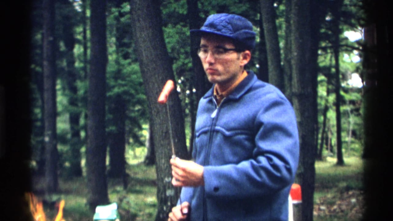 Man in blue cap and jacket roasts a hot dog on a stick in a wooded campsite.